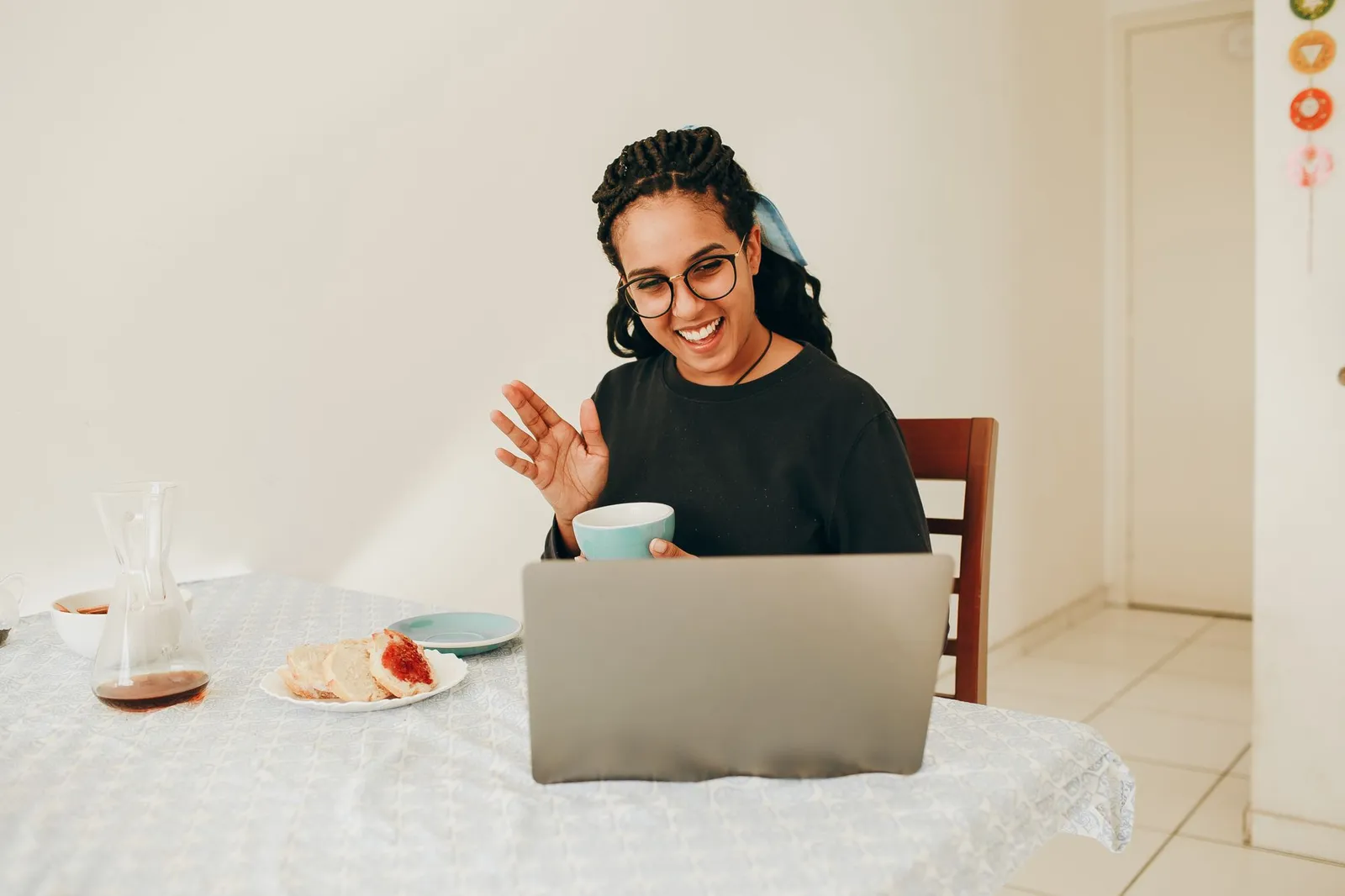 A smiling woman in glasses sits at a kitchen table at home, holding a cup and waving at the screen of an open laptop