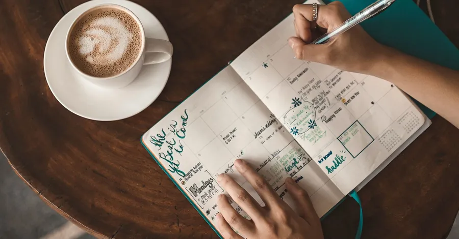 Overhead view of hands writing in an open monthly planner on a round wooden table, beside a cup of coffee on a saucer