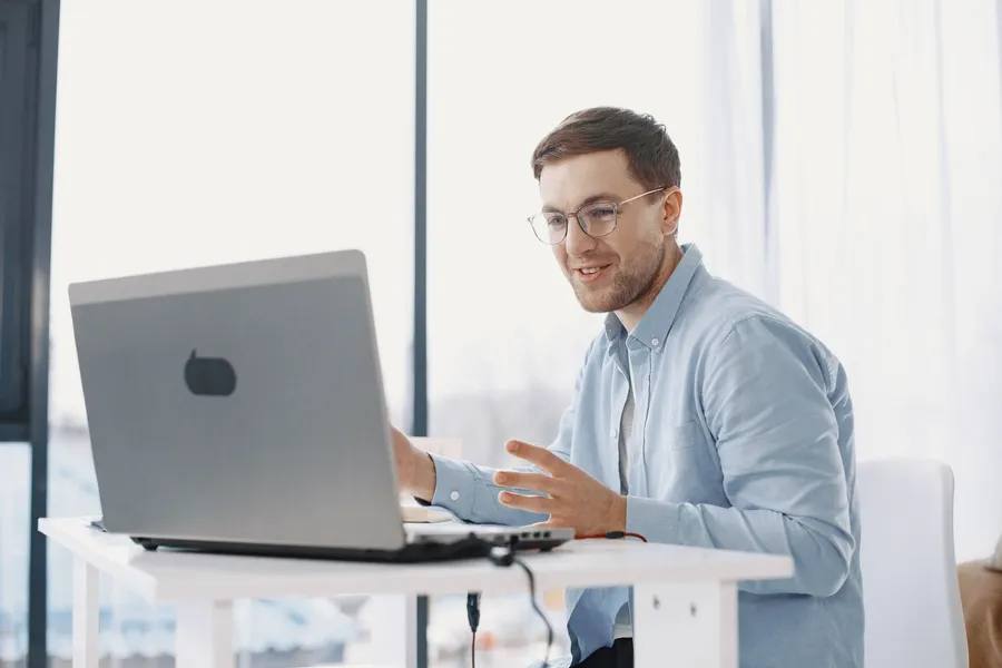 A man smiling while talking on a laptop video call by a window